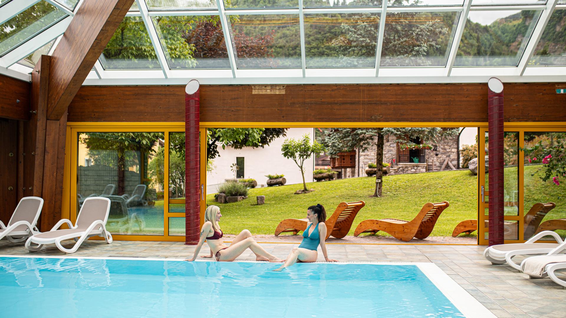 Due persone rilassate in piscina coperta, circondate da natura e tranquillità.