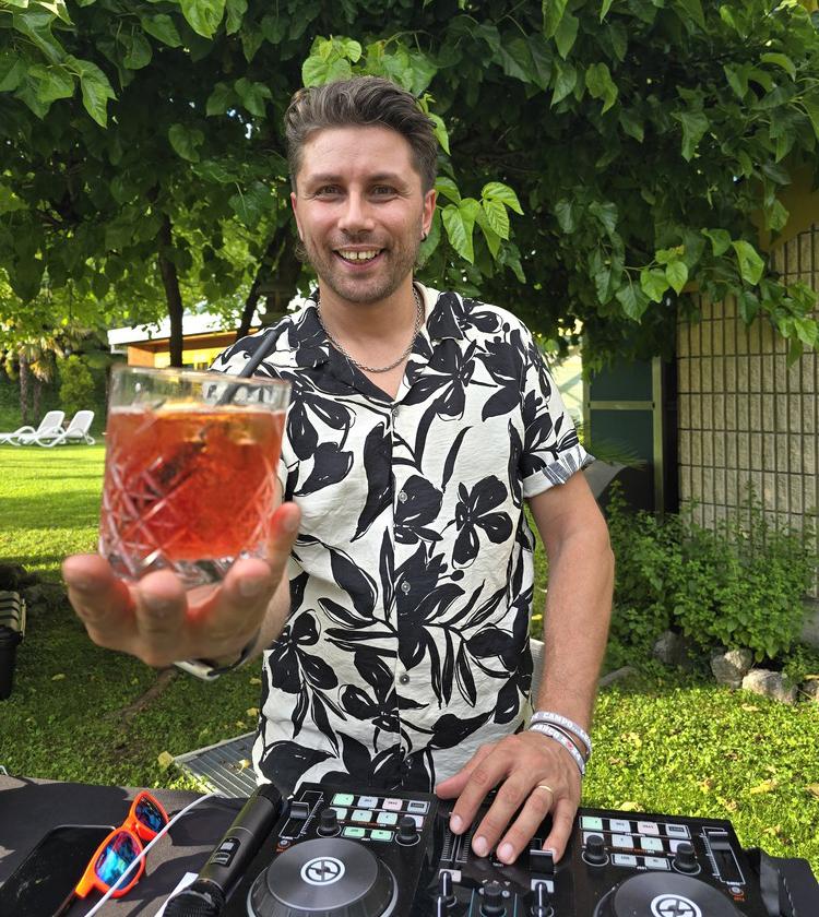 Uomo sorridente offre cocktail in giardino, atmosfera estiva e rilassata con musica in sottofondo.