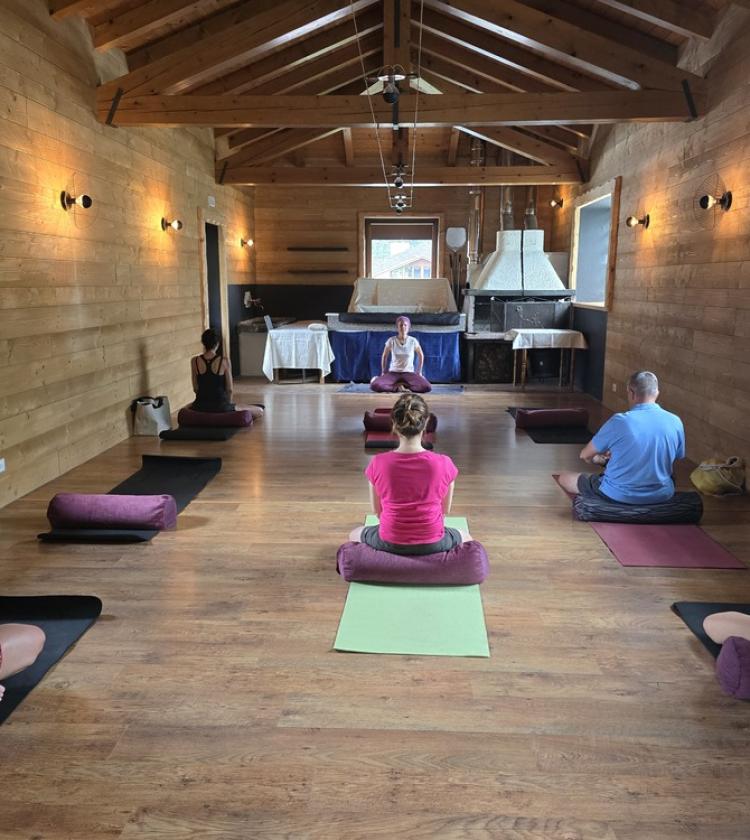 Meditazione in sala rustica, atmosfera serena con persone sedute su tappetini.