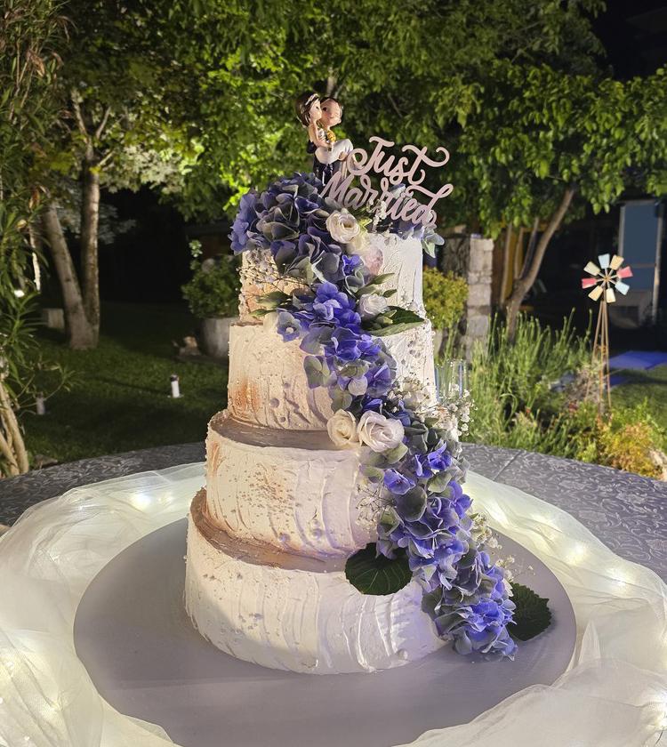 Torta nuziale a tre piani, decorata con fiori blu, statuetta e scritta 'Just Married'.